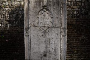 "Una lastra di pietra antica incastonata in una parete di mattoni e pietre rustiche. Al centro, la lastra presenta uno stemma araldico scolpito in rilievo nella parte superiore e un'iscrizione in latino, parzialmente usurata dal tempo, che ne occupa la sezione centrale."