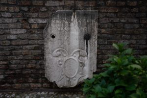 "Dettaglio di una facciata in intonaco chiaro, con segni di usura, che mostra uno stemma araldico dipinto tra due finestre. Lo stemma raffigura un leone dorato su sfondo grigio nella parte superiore e bande orizzontali rosse e bianche in quella inferiore. Ai lati si trovano due finestre con persiane in legno verde visibilmente scrostate dal tempo."