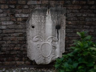 "Dettaglio di una facciata in intonaco chiaro, con segni di usura, che mostra uno stemma araldico dipinto tra due finestre. Lo stemma raffigura un leone dorato su sfondo grigio nella parte superiore e bande orizzontali rosse e bianche in quella inferiore. Ai lati si trovano due finestre con persiane in legno verde visibilmente scrostate dal tempo."