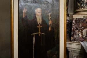 "Dipinto a olio raffigurante un santo barbuto con aureola, che indossa un saio scuro tipico degli ordini monastici, con un crocifisso al collo e un cordone annodato in vita. Il soggetto tiene la mano destra alzata con l'indice puntato verso l'alto e regge un piccolo crocifisso nella mano sinistra. Il quadro &egrave; inserito in una cornice dorata all'interno di un ambiente religioso."