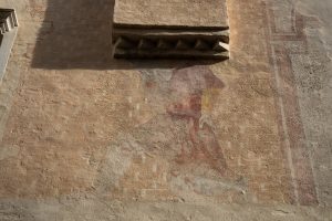 L'immagine mostra un dettaglio architettonico di una parete in intonaco color ocra/beige. Sotto una cornice in rilievo caratterizzata da una serie di modanature triangolari (a dentelli), si intravedono i resti di un affresco sbiadito che raffigura una figura umana, probabilmente un Santo o una figura allegorica, con vesti dai toni rossastri.