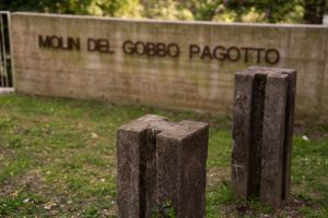 L'immagine mostra un'area all'aperto, probabilmente un parco o un sito storico, con una parete di fondo in calcestruzzo su cui sono fissate le lettere metalliche che compongono il nome. In primo piano si trovano due pilastrini in pietra o cemento di forma rettangolare, caratterizzati da una fessura verticale profonda che corre lungo il centro di ciascuna faccia.