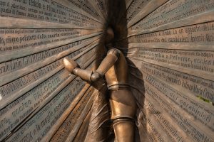 "Scultura in bronzo che raffigura una figura umana stilizzata, simile a un manichino articolato, posizionata al centro di una struttura formata da pannelli metallici che si irradiano verso l'esterno. Ogni pannello &egrave; ricoperto di testi incisi in rilievo che riportano cronache di eventi drammatici della storia italiana recente, tra cui attentati terroristici, omicidi di mafia e atti di violenza politica."