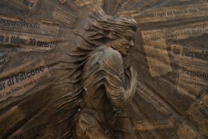 L'immagine mostra un monumento scultoreo in bronzo, caratterizzato da una figura umana centrale in rilievo, i cui tratti e vesti sembrano fondersi in un movimento dinamico. Lo sfondo della scultura &egrave; composto da numerosi riquadri che riproducono fedelmente titoli e articoli di giornale italiani, trasformando la superficie in una sorta di archivio storico inciso.