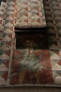 Dettaglio di una parete interna affrescata del Palazzo Marone a Pordenone. Al centro, un busto di guerriero medievale &egrave; raffigurato con un'armatura a scaglie verde, una spada alla cintura e un piccolo scudo circolare. Il guerriero ha una lunga barba bionda e uno sguardo fisso. Sopra di lui, una porzione di muro con un motivo geometrico a zig-zag e a rombi si erge come una colonna. Lo sfondo &egrave; rosso mattone e presenta segni di usura, mentre una serie di cornici dipinte circonda l'affresco.