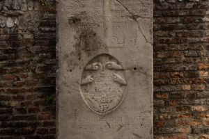 L'immagine mostra una lapide commemorativa o una pietra tombale in pietra chiara, posizionata tra due pareti di mattoni rossi. Al centro della lastra si trova un emblema araldico scolpito in bassorilievo all'interno di una forma ovale, raffigurante dei motivi floreali o animali stilizzati sormontati da una corona.