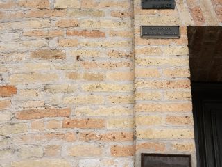 Questa fotografia ravvicinata inquadra l'angolo destro di un muro esterno in mattoni di terracotta di diverse tonalità, prevalentemente giallo-marrone e rosa, con evidenti segni del tempo e della malta grigia. A destra, si intravede una porta in legno scuro. Sul montante destro della porta sono fissate verticalmente tre targhe commemorative di metallo scuro, posizionate a diverse altezze. La targa superiore, più piccola e di forma rettangolare orizzontale, riporta l'iscrizione: "3 . 9 . 1985 / Mt. 13,05". La targa centrale, di dimensioni simili, recita: "26-11-2002 / Mt. 17,90 S.L.M.". La targa inferiore, più antica e di forma rettangolare orizzontale, presenta una cornice metallica più elaborata; l'iscrizione è incisa e include la data "28-10-1928" e l'elevazione "Mt. 17,90". Sopra la targa centrale, si vedono due bulloni metallici sporgenti dal muro, probabilmente parte di un ancoraggio rimosso o mai utilizzato. L'intero dettaglio riflette la storia e l'evoluzione dell'edificio nel corso del tempo.