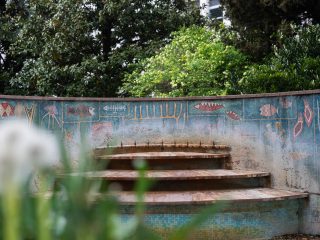 Gradini semicircolari in pietra e mosaico davanti a una parete decorata con simboli colorati; fiori e piante in primo piano e alberi sullo sfondo.