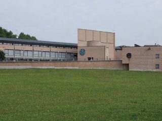 Veduta esterna panoramica del Centro Diocesano di Pordenone, sede del Museo Diocesano di Arte Sacra. L'edificio moderno in mattoni a vista presenta geometrie rigorose e un'ampia facciata con vetrate continue, situato davanti a un vasto prato verde sotto un cielo nuvoloso.