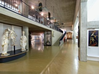 Una fotografia dell'interno del museo Diocesano d'arte sacra dallo spazio ampio, e pavimenti lucidi in resina beige e strutture in cemento armato a vista. Sulla sinistra, in primo piano, tre grandi statue lignee dipinte di bianco e oro sono esposte su una base scura. Al centro della sala, una rampa e una scala conducono a un ballatoio superiore protetto da una ringhiera metallica nera, dove altre sculture sono esposte su mensole di vetro sospese. Sulla destra, un pilastro in cemento ospita un dipinto incorniciato. In fondo alla sala si scorgono ulteriori opere d'arte, tra cui un altare ligneo dorato e una croce processionale. L'illuminazione è puntuale, con faretti che mettono in risalto i singoli pezzi esposti.