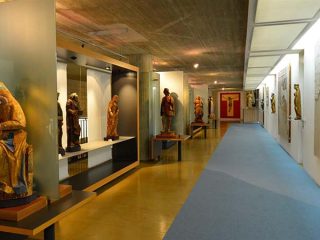 Fotografia del primo piano del museo Diocesano d'arte sacra, dedicato alla scultura lignea. Lungo il lato sinistro, diverse statue policrome di santi e Madonne con Bambino sono esposte su basi nere e separate da pannelli in vetro satinato. Sulla destra, un lungo corridoio con una passatoia azzurra è affiancato da una parete bianca su cui sono montati frammenti di affreschi e sculture a rilievo. In fondo al corridoio spicca un grande crocifisso su fondo rosso. Il soffitto in cemento grezzo e l'illuminazione a binario conferiscono all'ambiente un carattere moderno che contrasta con l'antichità delle opere esposte.