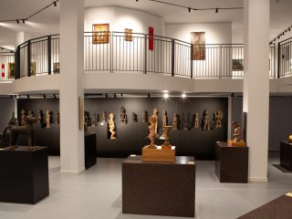 Fotografia della sala del museo Diocesano d'arte sacra dedicata alle mostre temporanee. Riprende una vasta collezione di sculture etnografiche e sacre. In primo piano, diverse statue sono esposte su solidi piedistalli scuri e quadrati, tra cui spicca sulla sinistra la figura di un felino e al centro una piccola statua di putto. Sullo sfondo, una parete scura ospita una lunga fila di statuette antropomorfe in legno e altri materiali, disposte su diversi livelli. L'architettura della sala è moderna, con colonne bianche che sostengono un ballatoio superiore circondato da una ringhiera metallica nera. Al piano superiore sono visibili quadri incorniciati e altre opere d'arte. L'illuminazione a binario crea un ambiente soffuso ma focalizzato sui reperti esposti, sottolineando il contrasto tra il bianco delle strutture e lo scuro delle basi espositive.