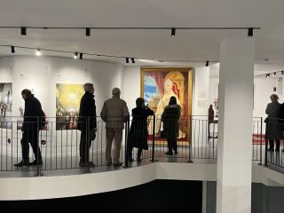 Fotografia del piano superiore della sala del museo Diocesano d'arte sacra dedicata alle mostre temporanee. Diverse persone sono affacciate a un ballatoio curvo, protetto da una ringhiera metallica grigia, mentre osservano le opere esposte. Al centro della parete di fronte, spicca un grande ritratto solenne in una cornice dorata che raffigura un pontefice (Papa Benedetto XVI) in abiti cerimoniali bianchi, seduto su un trono. L'ambiente è caratterizzato da grandi pilastri bianchi e da un sistema di illuminazione a binario sul soffitto che proietta luce puntuale sulle opere. Sul lato destro, altre persone osservano oggetti più piccoli e tappeti esposti in una zona adiacente.