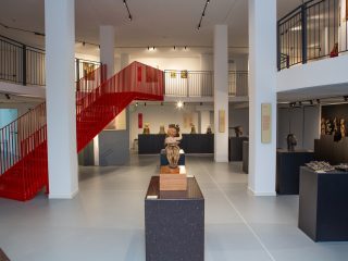 Una fotografia della Sala del museo Diocesano d'arte sacra dedicata alle mostre temporanee. L'elemento architettonico più distintivo è una scala metallica rosso acceso che risalta contro le pareti e i pilastri bianchi, conducendo al piano superiore. Al piano terra, diverse opere sono esposte su basamenti scuri e squadrati: in primo piano una scultura dorata, mentre sullo sfondo e sulla destra si notano campane antiche e una collezione di statuette etnografiche. L'ambiente è caratterizzato da un ballatoio superiore con ringhiere nere che si affaccia sulla sala centrale. L'illuminazione a binario e il pavimento grigio chiaro contribuiscono a creare un'atmosfera moderna e ordinata, mettendo in risalto il contrasto cromatico tra il bianco della struttura e il rosso della scala.