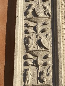 Rilievo decorativo in pietra con motivi vegetali e animali scolpiti, disposto verticalmente su una facciata.