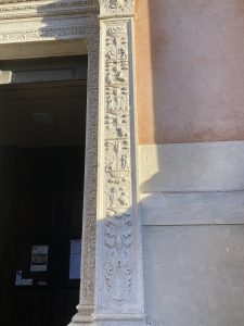 Dettaglio di uno stipite in pietra scolpita di un portale monumentale, riccamente decorato con bassorilievi in stile rinascimentale che presentano motivi a candelabra. La decorazione alterna elementi vegetali, come vasi e foglie d'acanto, a piccole figure umane e animali disposte in riquadri verticali. Il pilastro in pietra chiara contrasta con la parete adiacente di color rosa antico, parzialmente illuminata dalla luce solare laterale che ne esalta la tridimensionalit&agrave;. Sulla sinistra si intravede l'ingresso scuro di un edificio sacro con alcuni avvisi affissi sulla porta.