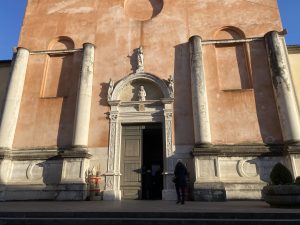 Facciata di una chiesa storica, caratterizzata da un intonaco color rosa tenue. L'ingresso principale &egrave; incorniciato da un portale in pietra lavorata, sormontato da una lunetta scolpita che raffigura Cristo, e coronato da una statua su una nicchia superiore. Due grandi colonne in pietra sorreggono idealmente la parte centrale della facciata, che presenta nicchie laterali vuote. Una persona &egrave; ferma sulla soglia del portone aperto, pronta a entrare. Ai lati del portale si notano decorazioni scultoree e vasi con piante. La luce del sole proietta ombre nette sulla struttura, mettendo in risalto i volumi architettonici.