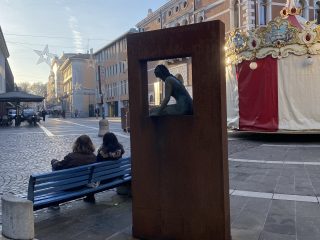 "Una veduta di Piazza Cavour a Pordenone in una giornata di sole. In primo piano, una scultura in bronzo raffigurante una figura femminile seduta è incastonata in un'alta struttura metallica rettangolare color ruggine. A sinistra della scultura, due persone sono sedute su una panchina blu, mentre sulla destra si intravede la parte di una giostra con tendaggi rossi e bianchi. Sullo sfondo si estende la piazza pavimentata con edifici storici e decorazioni natalizie a forma di stella sospese tra le strade."