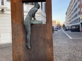 "Vista laterale di una scultura in bronzo situata in Piazza Cavour a Pordenone. L'opera raffigura una figura femminile che sembra scavalcare o essere seduta sul bordo di un'alta struttura metallica rettangolare color ruggine. Il corpo della scultura, lavorato con una texture a rilievo, è proteso in avanti, mentre una gamba pende lungo il lato esterno della cornice. Sullo sfondo, si vede una strada pavimentata che conduce verso il centro della piazza, fiancheggiata da eleganti edifici storici dai toni chiari e prospettive urbane."