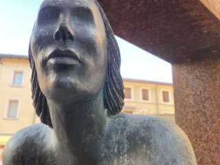"Primo piano ravvicinato del volto e delle spalle di una scultura in bronzo posta in Piazza Cavour a Pordenone. Il soggetto è una figura femminile con i capelli sciolti e il viso rivolto verso l'alto in un'espressione contemplativa. La superficie del bronzo presenta una patina scura con dettagli lavorati a rilievo e striature orizzontali sulla parte inferiore del busto. La statua è parzialmente incorniciata da una struttura metallica verticale dalla texture arrugginita, con sullo sfondo, leggermente sfuocati, gli edifici color ocra tipici della piazza."