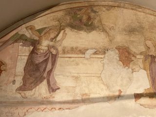 Dettaglio dell'affresco rinascimentale dell'Annunciazione dipinto all'interno di una lunetta raffigurante l'Angelo Gabriele con ali colorate che avanza verso la Vergine Maria, situata a destra in posa devota davanti a un leggio. In alto, Dio Padre emerge dalle nuvole e il Bambin Gesù vola verso la Vergine. L'opera presenta ampie lacune di intonaco bianco che interrompono la scena.