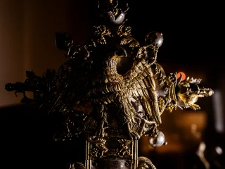 Un'inquadratura ravvicinata del terminale superiore di una croce processionale antica in metallo sbalzato e dorato. Al centro risalta la figura di un'aquila ad ali spiegate, simbolo dell'Evangelista Giovanni, resa con un minuzioso dettaglio del piumaggio. L'aquila poggia su una base decorata all'interno di una cornice polilobata, ornata da motivi floreali, viticci e piccole sfere metalliche. La luce radente proviene da destra, creando profondi contrasti d'ombra che enfatizzano la tridimensionalità della lavorazione orafa. Lo sfondo è scuro e indistinto, con un piccolo punto di luce calda che appare in lontananza.