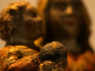 Un primo piano ravvicinato e con una profondità di campo ridotta di un'antica scultura religiosa in legno policromo. In primo piano Gesù Bambino tiene in mano un piccolo globo scuro; la vernice è visibilmente crepata e usurata dal tempo, rivelando gli strati sottostanti. Sullo sfondo, fuori fuoco, si scorge il volto della Madonna con lunghi capelli. L'atmosfera è calda, con un'illuminazione soffusa che enfatizza le texture materiche della statua.