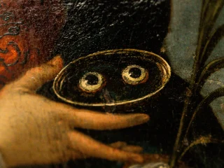Dettaglio di un dipinto antico che raffigura una mano che regge un piccolo vassoio contenente due occhi umani: gli occhi di Santa Lucia. Un'altra mano, in basso a destra, impugna una foglia di palma, simbolo di martirio. La pittura presenta una texture granulosa e scura, tipica delle tele antiche, con un forte contrasto tra i toni bruni dello sfondo e i dettagli illuminati.