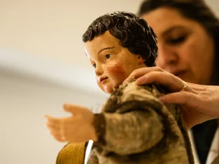 Primo piano di una statua lignea policroma di Gesù Bambino, con capelli ricci scuri e guance rosate. Una restauratrice, parzialmente visibile e sfocata sullo sfondo, poggia delicatamente le mani sulle spalle della scultura per stabilizzarla. Il Bambino indossa una veste antica in tessuto pesante e usurato. L'illuminazione è soffusa e calda, creando un'atmosfera di estrema cura e rispetto per il manufatto.