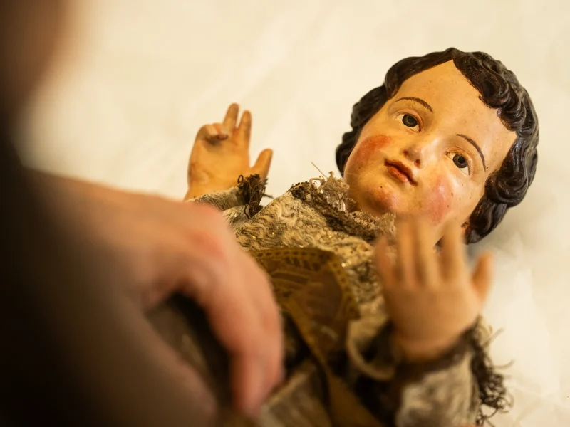 Inquadratura dall'alto di una statua lignea policroma raffigurante Gesù Bambino, distesa su una superficie bianca protettiva. Le mani della restauratrice al lavoro, parzialmente visibili in primo piano, sostengono con cura il busto della scultura. Il Bambino ha capelli scuri ricci, guance arrossate e indossa una veste in tessuto antico ricamato e usurato, con la mano destra sollevata in segno di benedizione.