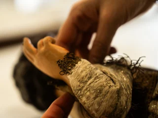 Primo piano macro delle mani di un restauratore che maneggiano con cura il polso di una statua lignea o in cera. La statua indossa una manica di tessuto antico pregiato, con trame argentate e bordi sfrangiati. Il gesto sottolinea la fragilità del manufatto e la precisione millimetrica necessaria per sistemare i paramenti storici senza danneggiarli.