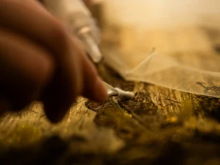 Inquadratura macro e dinamica delle mani di una restauratrice mentre pulisce con estrema precisione un antico tessuto dorato. Viene utilizzato un bastoncino con punta in cotone per agire sulle fibre delicate. Accanto alla zona di intervento è steso un velo protettivo trasparente. La luce calda mette in risalto la lucentezza dei filati metallici e la meticolosità del lavoro.