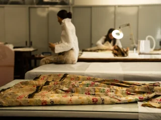 Interno del laboratorio di restauro tessile del museo Diocesano. In primo piano, un antico tessuto damascato con piccoli ricami floreali rossi e verdi è disteso su un tavolo da lavoro. Sullo sfondo, la restauratrice lavora con precisione sotto la luce di una lampada professionale, mentre un manichino con indosso un abito storico bianco è seduto su un altro tavolo, creando un'atmosfera di studio e conservazione artigianale.