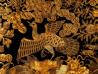 Dettaglio di un prezioso paramento sacro ricamato in oro su fondo scuro. Al centro spicca la figura di un uccello mitologico dalle ali spiegate, circondato da motivi ornamentali a nuvola e decorazioni floreali realizzate con fili dorati a rilievo.