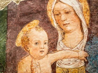 Un affresco rinascimentale del Tolmezzo raffigurante la Madonna col Bambino. Maria è rappresentata sulla destra, con il volto inclinato verso il figlio e uno sguardo dolce e pensieroso; indossa un velo bianco che le incornicia il viso e un'aureola dorata appena accennata. Il Bambino, in primo piano sulla sinistra, guarda verso l'osservatore con grandi occhi espressivi e la mano destra sollevata in un gesto di benedizione. L'opera mostra i segni del tempo, con una trama di sottili crepe (craquelé) sulla superficie e una tavolozza di colori caldi e terrosi, con sfumature di ocra, rosso bruno e azzurro polveroso sulle vesti. Il fondo è scuro e materico, mettendo in risalto la delicatezza del disegno dei volti.