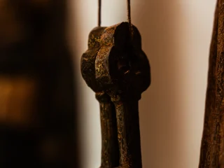 Un'inquadratura ravvicinata e frontale delle chiavi di San Pietro, parte di una scultura lignea antica. Due grandi chiavi in legno scuro sono appese verticalmente, incrociandosi leggermente. I fusti delle chiavi sono solidi e terminano con ingegni dalla forma complessa e geometrica. Il legno presenta una texture scabra e irregolare, con sfumature che vanno dal bruno scuro al ruggine. Lo sfondo è composto dalla superficie del legno scolpito della statua, con venature verticali profonde e residui di policromia dorata e verdastra, lasciata volutamente fuori fuoco per far risaltare il dettaglio delle chiavi.