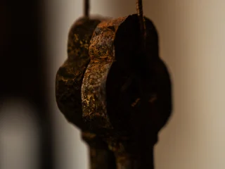 Un'inquadratura ravvicinata e artistica dell'impugnatura di due grandi chiavi antiche in metallo scuro, simbolo iconografico di San Pietro. Le chiavi sono appese a una sottile cordicella verticale e mostrano una superficie irregolare, segnata dall'ossidazione e dal tempo, con piccoli riflessi dorati che emergono dal metallo brunito. L'impugnatura ha una forma lobata classica. La profondità di campo è molto ridotta, lasciando lo sfondo completamente sfocato in tonalità neutre e calde, concentrando tutta l'attenzione sulla texture materica e sulla silhouette solida dell'oggetto.