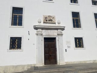 "Facciata di un edificio storico intonacato di bianco con un importante portale d'ingresso in pietra, sormontato da un bassorilievo del Leone di San Marco alato e due elementi decorativi a forma di fiamma. Sopra la porta in legno scuro è incisa l'iscrizione 'MONS PIETATIS A FVNDAMENTIS ERECTUS MDCCLXVII'. La facciata presenta finestre simmetriche protette da grate in ferro battuto; a destra dell'ingresso è visibile una targa che riporta le scritte 'Biblioteca Civica' e 'Casa della Musica'."
