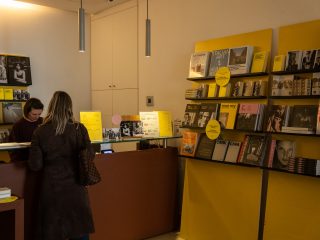 Una fotografia dell'interno di un bookshop o di una biglietteria di un museo, caratterizzata da pareti bianche e scaffalature giallo acceso. Sulla destra, numerosi libri d'arte e di fotografia (con nomi visibili come Man Ray e Capa) sono esposti su mensole nere fissate a pannelli gialli. Al centro, una cliente di spalle con un cappotto marrone e una borsa a tracolla interagisce con una dipendente dietro un bancone in legno scuro. Sul bancone sono presenti listini prezzi e piccoli espositori. L'ambiente è illuminato da sottili lampade a sospensione cilindriche dal design moderno.