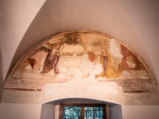 Fotografia di un affresco frammentario raffigurante l'Annunciazione, situato all'interno di una lunetta sotto una volta a crociera bianca. L'opera mostra l'Arcangelo Gabriele a sinistra e la Vergine Maria a destra, con Dio Padre e il Bambin Gesù in alto al centro. Sotto l'affresco si apre una finestra che si affaccia su del fogliame verde. La struttura architettonica circostante è sobria e intonacata di bianco.