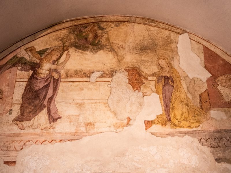 Affresco rinascimentale dell'Annunciazione dipinto all'interno di una lunetta. A sinistra, l'angelo Gabriele con ali colorate avanza verso la Vergine Maria, situata a destra in posa devota davanti a un leggio. Al centro in alto, Dio Padre emerge dalle nuvole e il Bambin Gesù vola verso la Vergine. L'opera presenta ampie lacune di intonaco bianco che interrompono la scena, in particolare tra le due figure e lungo il bordo inferiore.