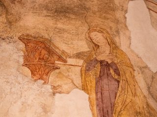 Dettaglio di un affresco parzialmente conservato che raffigura la Vergine Maria in preghiera. La figura ha il capo inclinato, le mani incrociate sul petto e indossa una veste viola coperta da un mantello ocra. Alla sua sinistra si trova un leggio in legno decorato con la scultura di un leone alato. L'intonaco originale è frammentario, con bordi irregolari che rivelano la muratura retrostante.