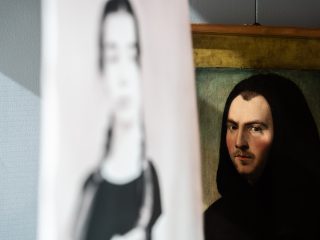 Un gioco di prospettive e riflessi all'interno di una sala museale del Ricchieri. In primo piano, una sagoma sfocata e semitrasparente di una figura femminile contemporanea della fotografa Olivia Arthur sembra sovrapporsi a un ritratto classico ad olio posizionato sullo sfondo. Il dipinto ritrae un monaco con un cappuccio del saio che gli copre il capo e uno sguardo intenso rivolto verso l'osservatore. Il contrasto tra l'antico e il contemporaneo crea un effetto etereo e senza tempo.