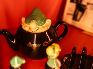 Un primo piano dettagliato della teiera di Andy Capp già vista nell'immagine precedente. La teiera, realizzata in ceramica nera lucida, presenta la scritta dorata "ANDY CAPP" sul corpo bombato. Il coperchio è l'elemento più caratteristico: riproduce il volto sorridente del personaggio dei fumetti, con il suo iconico berretto a scacchi verde calato sugli occhi. Sullo sfondo, sfocato ma visibile, c'è il resto della collezione: In basso si vedono le teste delle piccole statuine di Andy e Flo. Sulla destra compare parzialmente il porta-toast nero. Lo sfondo rosso intenso e l'illuminazione calda mettono in risalto la finitura lucida della ceramica, sottolineando la natura collezionistica dell'oggetto.