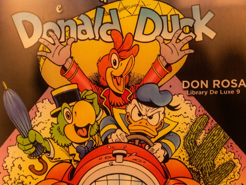 Una vivace illustrazione a fumetti che funge da copertina per un volume intitolato "Donald Duck" di Don Rosa (Library De Luxe 9). La scena, dinamica e colorata, mostra tre celebri personaggi Disney a bordo di un'auto rossa scoperta: Al centro: Paperino è alla guida con un'espressione decisa e un po' accigliata, le mani strette sul volante. Indossa la sua classica casacca da marinaio blu e gialla. A sinistra: José Carioca, il pappagallo verde, sorride allegramente mentre tiene un ombrello chiuso in mano. Indossa un elegante abito giallo, un papillon blu e il suo tipico cappello di paglia. Sullo sfondo: Panchito Pistoles, il gallo rosso, balza in piedi dietro i compagni con le ali spalancate in un gesto esultante e un'espressione di pura gioia. I tre personaggi sono incorniciati da una nuvola di polvere bianca che suggerisce l'alta velocità dell'auto, ambientata in un paesaggio desertico accennato da un cactus sulla destra. Il titolo "Donald Duck" appare in grandi lettere azzurre e bianche nella parte superiore, sovrapposto a un grande sole giallo circolare. Lo stile del disegno è quello dettagliato e cinetico caratteristico dell'autore Don Rosa.