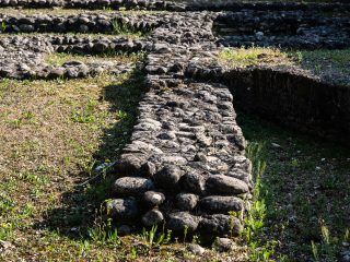 Veduta di dettaglio dell'area archeologica della villa romana di Torre che mostra i resti delle fondamenta di un settore dell'edificio. I muretti bassi sono realizzati con ciottoli di fiume legati da malta, circondati da erba rada e vegetazione boschiva sullo sfondo.