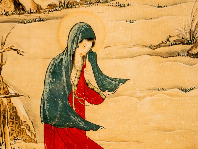 Un dettaglio a mezza figura di un dipinto a inchiostro e colore su seta in stile tradizionale dell'Asia orientale, raffigurante la Vergine Maria. La figura femminile è mostrata di trequarti, rivolta leggermente verso destra, con gli occhi abbassati e un'espressione serena e contemplativa. Indossa un mantello di colore teal scuro con sfumature verdastre e bordi decorativi, che le copre la testa e scende sulle spalle e sulle braccia, fluttuando leggermente all'estremità destra. Sotto il mantello, indossa una veste rossa brillante, con un colletto e maniche che mostrano dettagli di tessuto più chiari e modellati, e un nastro verde sottile annodato in vita. Le sue mani sono giunte delicatamente davanti al petto. Intorno alla sua testa c'è un nimbus (aureola) circolare, di un giallo-arancio caldo e tenue, che si staglia contro lo sfondo. Lo sfondo è un paesaggio stilizzato di dune di sabbia dorata o neve, con elementi vegetali sparsi e linee ondulate che ne suggeriscono la texture. A sinistra, si intravede una struttura architettonica o un albero stilizzato e spoglio. A destra, un ciuffo d'erba più grande. L'intero dipinto ha una trama superficiale di tela di seta, con colori tenui e fluidi che mostrano segni di usura storica, come sbiadimenti e crepe, tipici dei dipinti orientali antichi.
