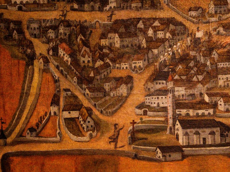 Parte di un'opera storica che rappresenta una veduta aerea del villaggio di Cimolais durante l'incendio del 1913. Il dipinto mostra una fitta rete di case con tetti spioventi e strade in terra battuta che convergono verso il centro. Sulla destra, spicca una chiesa bianca con un alto campanile dalla cuspide rossa. In primo piano, una figura in ginocchio prega davanti a una croce di legno. In alto a destra, fiamme e fumo si levano da alcuni edifici. I toni sono caldi, dominati dall'ocra, dal bruno e dal rossastro.