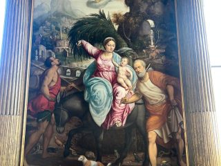 "Un dipinto di grandi dimensioni incorniciato tra due colonne classiche scanalate, opera di Pomponio Amalteo. Il quadro raffigura la Fuga in Egitto: la Vergine Maria, vestita con abiti rosa e azzurri, è seduta su un asino e tiene in braccio il Bambino Gesù, mentre solleva un grande ramo di palma. È accompagnata da San Giuseppe, che cammina a destra tenendo un bastone, e da un uomo di spalle sulla sinistra che guida l'asino. La scena è ambientata in un paesaggio ricco di dettagli naturali, inclusi vari animali (un cane, un coniglio, uccelli) in primo piano, formazioni rocciose e un borgo sullo sfondo, sotto il volo di un angelo."