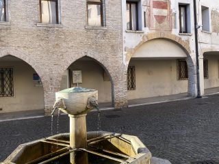 "Veduta di una piazza storica pavimentata in acciottolato. In primo piano si trova una fontana ottagonale in pietra con una vasca centrale dotata di mascheroni a forma di testa di leone da cui scorre l'acqua. Sullo sfondo, due edifici antichi presentano portici ad arco e facciate in mattoni e intonaco, con un affresco araldico visibile sulla parete del fabbricato a destra."